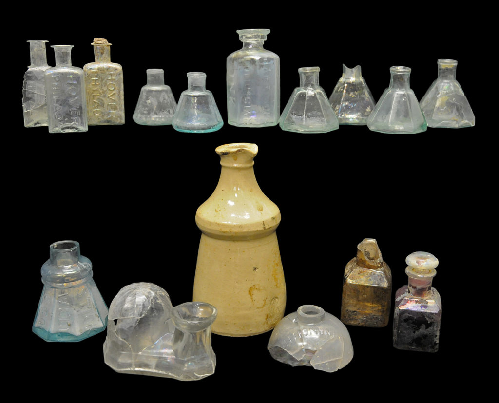 Assorted ink and mucilage bottles: (Top row, left to right) Two cone inks with molded ribs and label panels (4A-G-0105 and 4A-G-0038); “HARRISON’S / COLUMBIAN / INK” master ink bottle (4A-G-0325); four umbrella inks (4A-G-0335, Cat # 4.24.292, 4A-G-0336, 4A-G-0173). (Bottom row, left to right) “STICK / WELL / & CO” mucilage bottle (4A-G-0104); “MORGAN’S PATENT JULY 16TH 1867” (4A-G-0136); mucilage; stoneware master ink with impressed mark of Thaddeus Davids & Company stoneware master ink (4A-C-0038); domed ink embossed “UNION INK Co” (4A-G-0169) and two carmine inks (4A-G-0161 and 4A-G-0102).