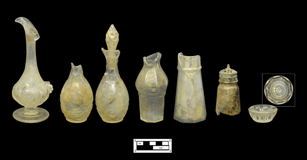 Colorless glass cruet, casters and salt (Left to right): This cruet and the two matching caster bottles were most likely used on the table during formal dining (4A-G-0132; 4A-G-0299; 4A-G-0300). The bottle in the center represents another style of caster, shaped with a wide shoulder to sit in a raised metal frame (4A-G-0281). The two conical shaker style bottles may have been used in the kitchen or on the table (4A-G-0167; Cat # 4.27.101). Two views of an individual pressed salt dish in the “Argus” pattern (4A-G-0330). 