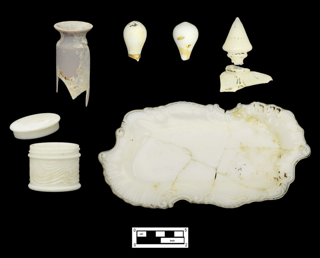 Sampling of fragrance, cosmetic and dresser containers: (Top row, left to right) Opaque amethyst bottle neck (Cat # 4.24.448); two opaque pale aqua fragrance bottle stoppers with remnants of gilt decoration (Cat # 4.24.446, 4.24.457); finial and lid fragment of translucent crackle glass ornamented with gilding and beaded enamel (Cat # 4.24.458, 4.27.121). (Bottom row, left to right) Opaque white lid from glass box (Cat # 4.24.463); opaque white glass “Larkin” jar (4A-G-0042); opaque white pressed glass dresser tray (4.24.475). 